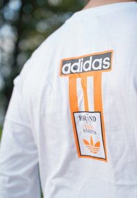 Λευκό μακρυμάνικο T-shirt με το μαύρο και πορτοκαλί λογότυπο της Adidas, έντονα γράμματα και λουλουδάτο γραφικό με το στοιχείο τριών ρίγων στη πλάτη.