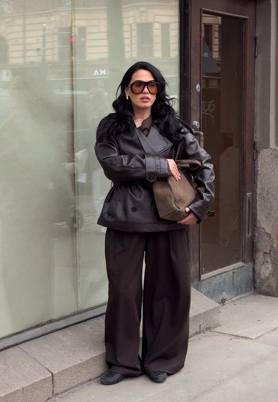 Mujer con cabello largo y negro, lleva gafas de sol grandes, chaqueta de cuero oscura, pantalones anchos, sostiene un bolso marrón y está parada en una acera de la ciudad.