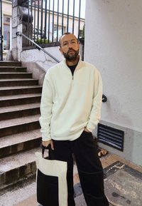 Fehér, zipzáras kapucnis pulóver, fekete nadrág, bézs és fekete táskát tart a kezében. Sima textúrájú, laza szabású, látható varrási részletekkel.