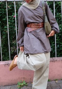Túnica cinza solta com um cinto largo de couro castanho, calças largas bege claro e sapatos slip-on de palha em tom de areia, acompanhados de uma bolsa verde de ombro.
