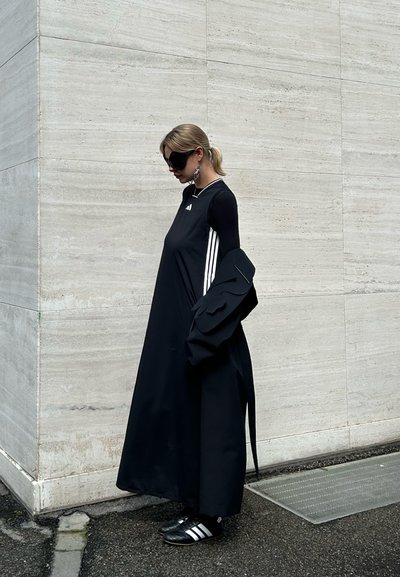 Vestido negro sin mangas con rayas blancas, superpuesto a una blusa de manga larga. Chaqueta negra con detalles texturizados y zapatos negros con acentos blancos.