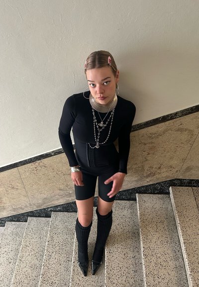 Body negro de manga larga combinado con pantalones cortos negros. Los accesorios incluyen collares metálicos en capas, pendientes grandes de aro y calcetas hasta la rodilla.