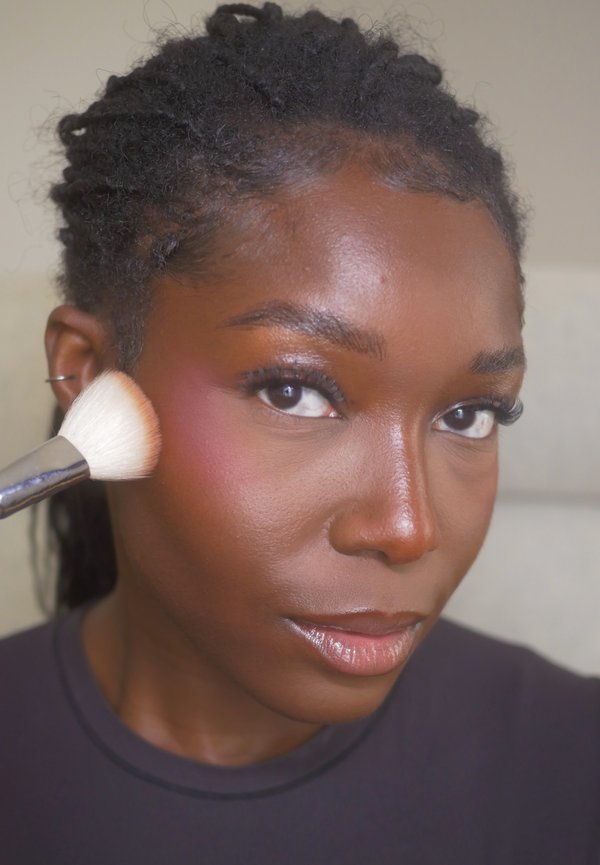 Personne à la peau foncée appliquant un blush rose sur la joue avec un pinceau à maquillage, regardant légèrement sur le côté.