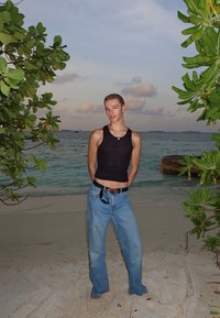 Juodas rankovių marškinėlis, šviesiai mėlynos plačios džinsinės kelnės ir juodas diržas su raudonu akcentu, stovintis paplūdimyje tarp žalumos.