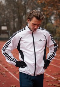 Veste de sport blanche avec accents noirs, fermeture éclair à l'avant, col montant, et trois rayures noires sur les manches. Portée avec des gants noirs et un pantalon bleu.