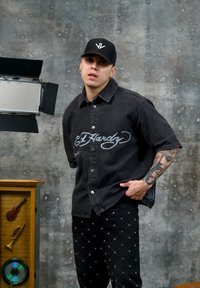 Jeune homme au bras tatoué portant un t-shirt noir Ed Hardy et une casquette, posant à l'intérieur contre un mur gris texturé avec une décoration vintage et un éclairage de studio.
