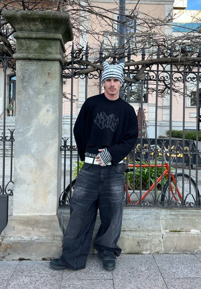 Sudadera negra recortada con un gráfico, jeans de mezclilla oscuros de pierna ancha, gorro de punto a rayas y cinturón blanco. De pie junto a un pilar de piedra y una cerca.