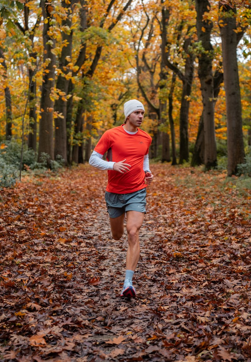 Runner wearing an orange short-sleeved shirt, grey shorts, and a white beanie, jogging along a leaf-strewn path in a forest adorned with autumn foliage.