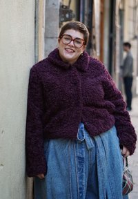 Personne aux cheveux courts, portant de grandes lunettes et un piercing au septum, souriant, vêtue d'un pull violet texturé et d'une jupe en denim bleu, debout près d'un mur.