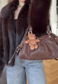 Femme portant une veste en fourrure marron foncé et un jean bleu clair, tenant un sac à main en cuir marron avec un porte-clés petit ours en peluche.