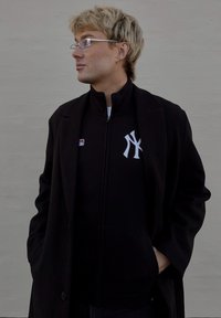 Černá bunda na zip s bílým logem New York Yankees, nošená pod černým kabátem. Outfit doplňují průhledné brýle a světlé vlasy.