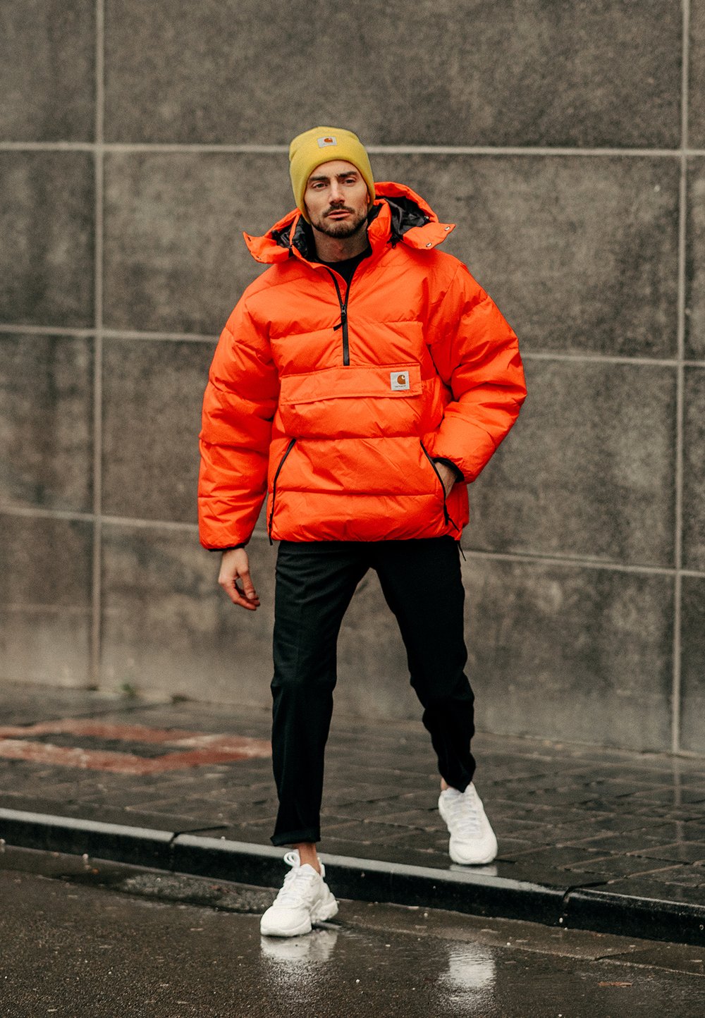 Casaco puffer laranja com fecho de ¼, apresentando um patch de logo e detalhes em preto. Combinado com calças pretas e ténis brancos.