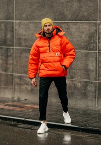 Casaco puffer laranja com fecho de ¼, apresentando um patch de logo e detalhes em preto. Combinado com calças pretas e ténis brancos.