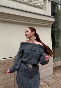 Pull en maille gris à épaules dénudées avec manches longues, texture côtelée, et ceinture noire ornée d'une boucle en métal, posé dans une rue.