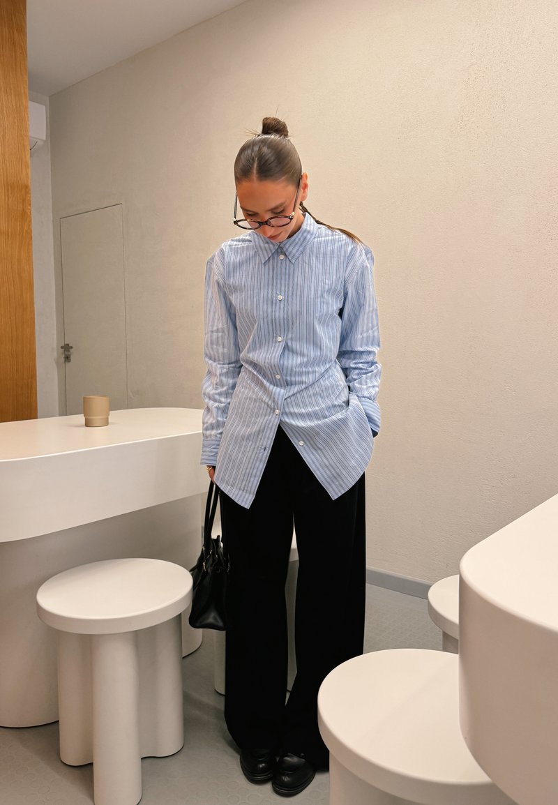 Hellblau gestreiftes Hemd mit Button-Down-Kragen, kombiniert mit schwarzen, weit geschnittenen Hosen. Die Accessoires umfassen eine schwarze Handtasche und eine Brille.
