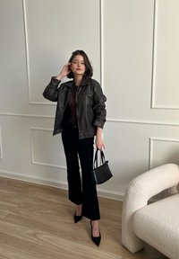 Barna bőrkabát, sötét felső, fekete, bő szárú nadrág és hegyes orrú fekete sarkú cipő. Egy kis fekete táskát tart, egy világos falnak támaszkodva.