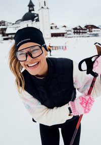 Νεαρή γυναίκα με εξοπλισμό για σκι χαμογελά, κρατώντας ένα μπαστούνι σκι σε χιονισμένη πλαγιά με χωριό και εκκλησία στο βάθος.