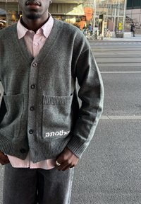 Sivi pleteni kardigan s V-izrezom, s gumbima na prednjem dijelu i dva džepa, s rebrastim manžetama i riječju "another" u bijeloj boji na rubu. Nosite ga preko ružičaste majice sa prugama.