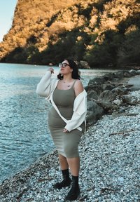 Vestito aderente verde oliva chiaro abbinato a un cardigan bianco a maglia grossa; stivali neri alla caviglia; in piedi vicino a una riva rocciosa con acqua.