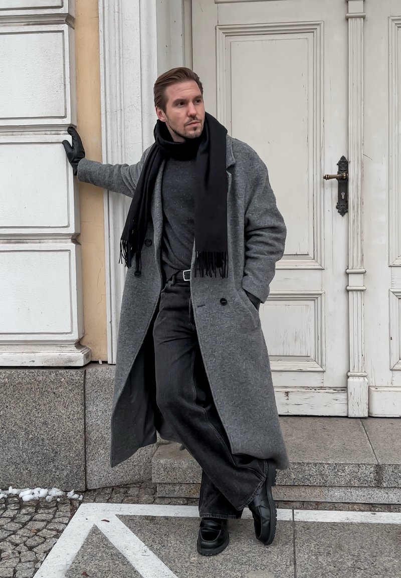 Grey wool overcoat, black scarf, black gloves, dark jumper, and dark jeans. Standing against a white door with detailed trim.