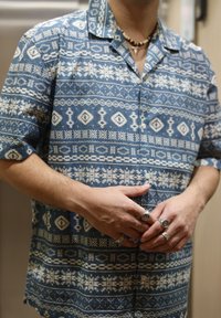 Chemise à manches courtes bleue avec des motifs géométriques blancs, un bouton sur le devant et un col. Portée avec plusieurs bagues en argent aux doigts.