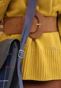 Pull en tricot jaune moutarde cintré par une large ceinture en daim marron, sangle bleu marine avec boucle, et sac à carreaux noirs partiellement visible.