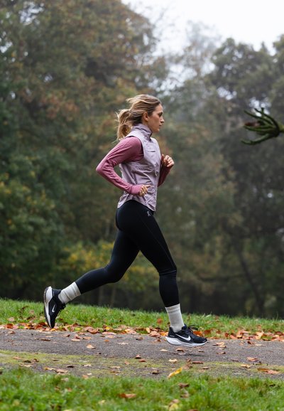 Una persona corre por un sendero, vistiendo un chaleco púrpura claro, una camiseta de manga larga color burdeos, leggings negros y zapatillas de correr negras con calcetines blancos.