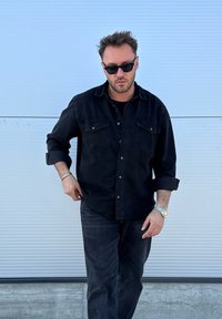 Čierna džínsová košeľa s dvomi náprsnými vreckami, rozopnutá pri golieri, v kombinácii s čiernymi džínsami. Doplnky zahŕňajú strieborné hodinky a slnečné okuliare.