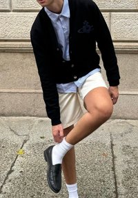 Cardigan noir en maille, chemise rayée blanche, shorts crème, chaussettes blanches et mocassins noirs. Jambes pliées, main touchant le genou, fond de rue en ville.