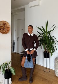 Vrouw staat binnen gekleed in een bruine gebreide trui, witte blouse, bruine rok, hoge bruine laarzen, en houdt een blauwe tas met studs vast.