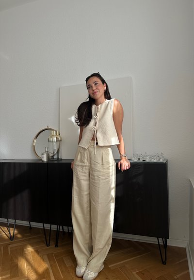 Chaleco de textura cremosa con cierre de lazo, combinado con pantalones de lino de pierna ancho en color beige. Suelo de madera clara y armario oscuro al fondo.
