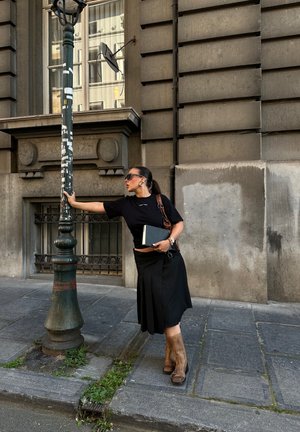 Camiseta negra y falda larga negra con pliegues, botas marrones hasta la rodilla, sosteniendo un cuaderno, junto a una farola pintada de verde.