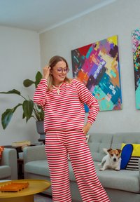 Rdeče in bele črtaste komplet loungewear, ki vključuje dolgo rokavičasto majico in hlače sproščenega kroja. Gladek material s sproščenim dizajnom in poleg ležeč pes.