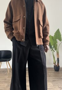 Casaco de lã castanho com dois bolsos frontais, camisa preta de botões e calças largas pretas. Ambiente interior com uma planta ao fundo.