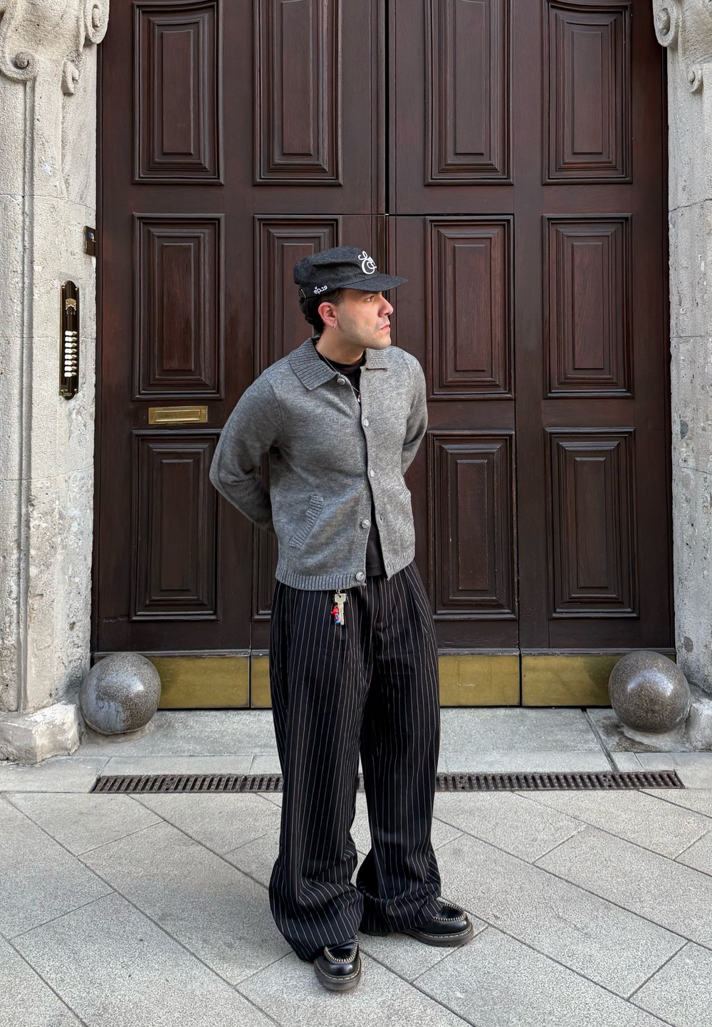 Uomo con giacca grigia, pantaloni neri a righe sottili e cappello, in piedi con le mani dietro la schiena davanti a grandi porte doppie in legno.