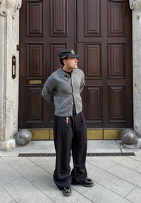 Uomo con giacca grigia, pantaloni neri a righe sottili e cappello, in piedi con le mani dietro la schiena davanti a grandi porte doppie in legno.