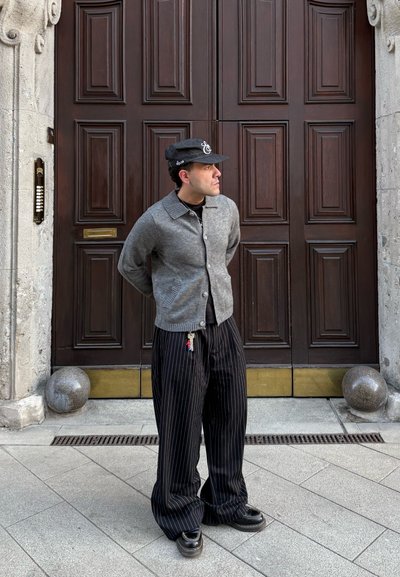 Hombre con chaqueta gris, pantalones negros de rayas y gorra, de pie con las manos detrás de la espalda frente a unas grandes puertas dobles de madera.