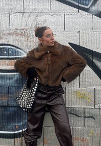 Καφέ ψεύτικη γούνα κοντό μπουφάν με ψηλό γιακά, μαύρο δερμάτινο παντελόνι και ένα μαύρο τσάντα με τρουκς. Το μοντέλο ποζάρει μπροστά από έναν υφασμάτινο τοίχο.