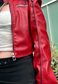 Jaqueta cropped em pele sintética vermelha com fecho éclair, apresentando detalhes em preto e texto bordado "Iconic Girl LTD. Edição de 1998" no peito.