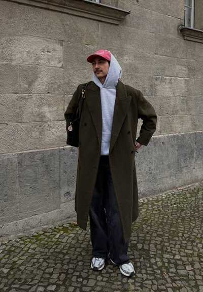 Abrigo de corte cruzado en verde oliva sobre una sudadera gris con capucha, bolso negro y jeans anchos oscuros. Zapatillas plateadas reflectantes completan el look.