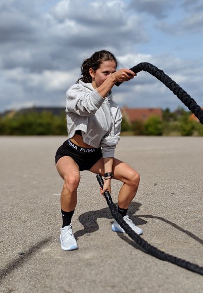 Mujer atlética con una sudadera gris recortada y pantalones cortos negros, usando cuerdas de batalla gruesas y negras. Lleva zapatillas blancas y un reloj negro.