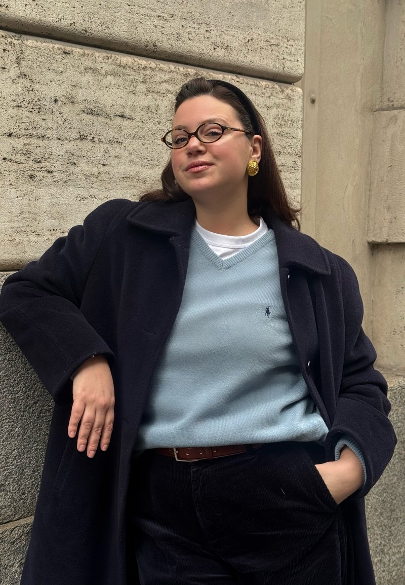 Mujer joven con cabello oscuro y gafas, apoyada en una pared de piedra, vestida con abrigo azul marino, suéter azul claro, pendientes dorados y pantalones negros.