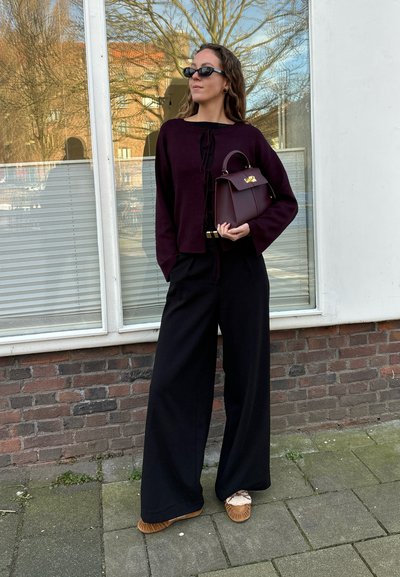 Suéter burdeos recortado con lazo delantero, pantalones anchos negros, zapatos de plataforma marrones y un pequeño bolso burdeos con herrajes dorados.