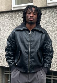 Czarna skórzana kurtka typu bomber z zamkiem błyskawicznym, ściągaczami przy mankietach i akcentami na ramionach. Noszona na szarych plisowanych spodniach. Teksturowane tło.