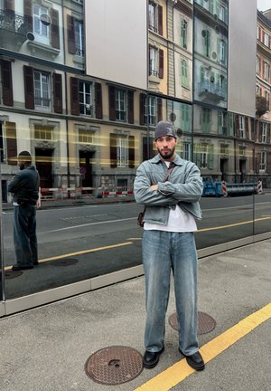 Hombre con una chaqueta de denim azul claro sobre una camisa blanca y jeans azules holgados, combinados con zapatos negros, de pie frente a un edificio con espejos.