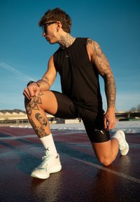 Αθλητής με τατουάζ, ντυμένος με μαύρα αθλητικά ρούχα, γονατισμένος σε μία πίστα, κοιτάει προς τα αριστερά, φοράει λευκά αθλητικά παπούτσια και γυαλιά ηλίου κάτω από καθαρό γαλανό ουρανό.