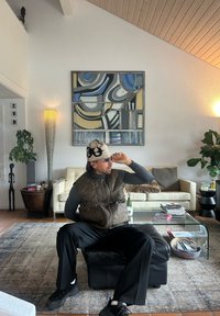 Polstret brun vest over en grå langermet skjorte, sittende på en svart skinnpuff. Beige hatt med svart mønster, briller og mørke bukser.