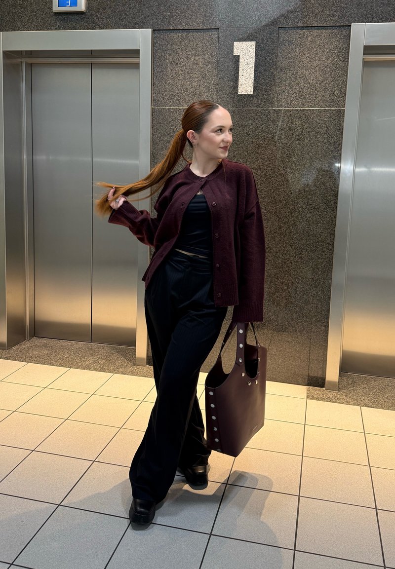 Femme avec queue de cheval portant des vêtements foncés tient un grand sac fourre-tout assorti, debout près de deux ascenseurs fermés et du numéro "1" sur le mur.