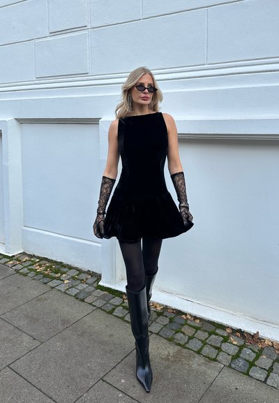 Vestido de terciopelo negro con falda acampanada, combinado con guantes de encaje y botas negras hasta la rodilla. Los accesorios incluyen unas pequeñas gafas de sol negras.