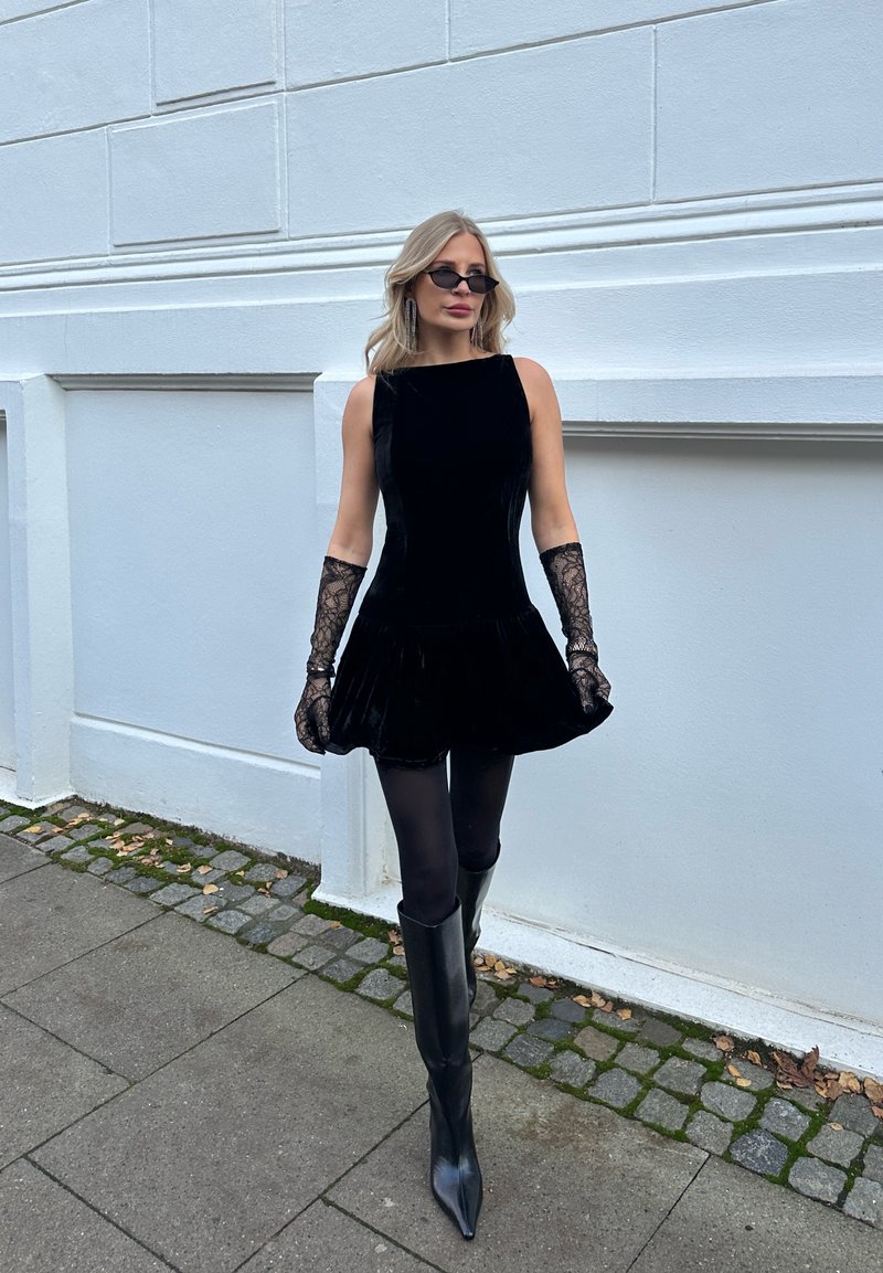 Robe en velours noir avec une jupe évasée, associée à des gants en dentelle et des bottes noires montant jusqu’au genou. Accessoires : petites lunettes de soleil noires.